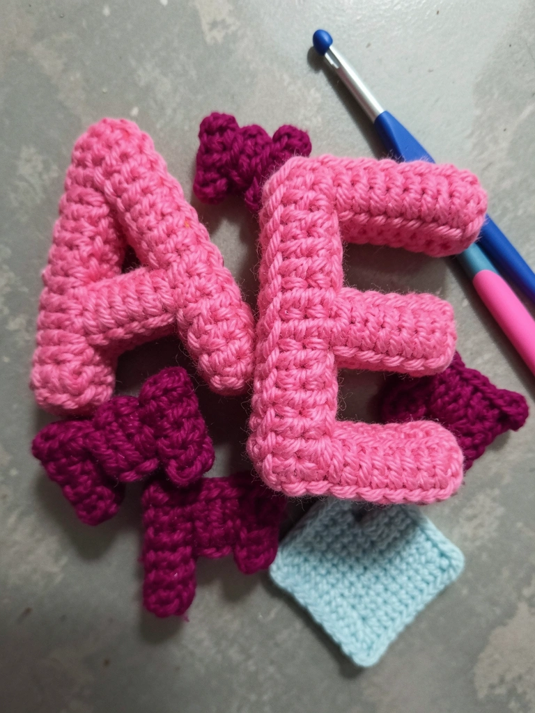Letras coloridas em amigurumi sobre mesa de madeira clara, com fios de lã e agulha ao lado